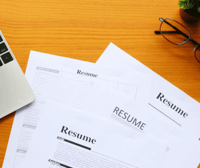 a stack of four resumes sits on a desk in between a laptop keyboard, a pair of glasses, and two small plants