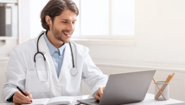 young male doctor in a lab coat with stethoscope over his shoulders smiles into laptop while writing notes